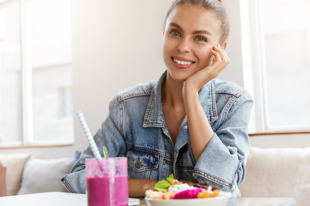 Positive female with attractive appearance has broad smile, dressed in denim jacket, spends free time at cafeteria, drinks smoothie and eats fruit salad, waits for friend. People and rest concept